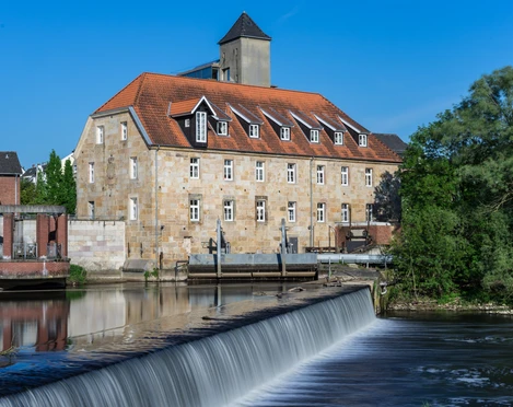 Rheine_Emswehr 2_Foto_ Rheine Tourismus Veranstaltungen eV_Y Mokdad.jpg Historisches Gebäude mit rotem Ziegeldach und Emswehr im Vordergrund bei sonnigem Wetter in Rheine.
