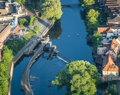 Rheine_Emswehr aus der Luft_Foto_ Rheine Tourismus Veranstaltungen eV.jpg Blick von oben auf das Emswehr in Rheine, umgeben von üppigem Grün und Gebäuden links und rechts.