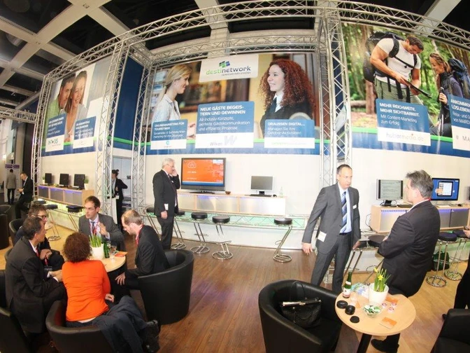 testbild Herzlich Willkommen auf einer Messe. Die Szene zeigt Menschen im Gespräch an einem Informationsstand.