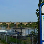 Anleger Sächsische Dampfschifffahrt Pirna Blick auf eine steinerne Brücke und den Anleger der Sächsischen Dampfschifffahrt über einen Fluss in Pirna, mit einem Informationsschild im Vordergrund und einem klaren Himmel.