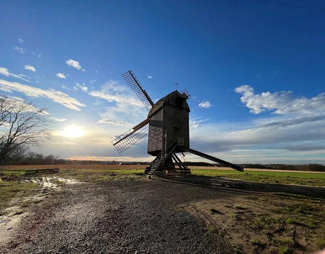 Mühle Neuenknick Historische, hölzerne Windmühle auf einem Feld bei Sonnenuntergang, umgeben von weitem Himmel und Wolken.