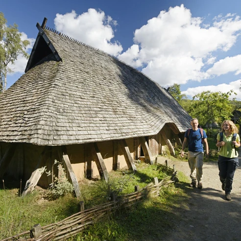 Rekonstruiertes Langhaus aus Holz mit Strohdach, zwei Personen spazieren auf einem Waldweg vorbei.