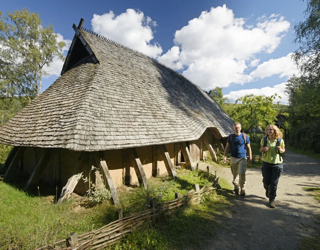 Oerlinghausen-AFM-Langhaus-Teutoburger-Wald-Tourismus-F-Grawe (50)_klein.jpg Rekonstruiertes Langhaus aus Holz mit Strohdach, zwei Personen spazieren auf einem Waldweg vorbei.