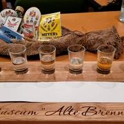 Korn Tasting - alte Brennerei Hille Sechs Schnapsgläser auf einem Holzkastl mit der Aufschrift "Museum Alte Brennerei", Tasting-Set.