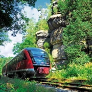 Nationalparkbahn U28 Ein roter Zug fährt durch eine grüne, bewaldete Landschaft mit großen Felsen im Hintergrund unter einem blauen Himmel.