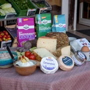 Das Bild zeigt eine liebevoll arrangierte Auswahl an biologischen Backwaren, verschiedenen Käsesorten und Müsli-Schalen, die appetitlich auf einem Tisch drapiert sind. Im Hintergrund sind frisches Bio-Gemüse und Obst zu sehen, die die Vielfalt und Frische des Sortiments unterstreichen.