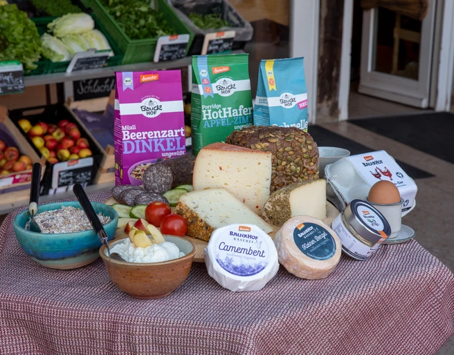 Bauckhof - Sortiment.jpg Das Bild zeigt eine liebevoll arrangierte Auswahl an biologischen Backwaren, verschiedenen Käsesorten und Müsli-Schalen, die appetitlich auf einem Tisch drapiert sind. Im Hintergrund sind frisches Bio-Gemüse und Obst zu sehen, die die Vielfalt und Frische des Sortiments unterstreichen.