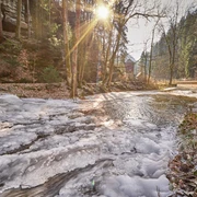 Flößersteig im Kirnitzschtal Verschneiter Bach in einem Wald bei Sonnenaufgang, mit Sonnenstrahlen durch die Bäume und einem kleinen Gebäude im Hintergrund.