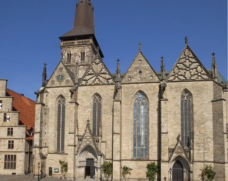 Außenansicht.jpg Eine historische Kirche mit markantem Turm, umgeben von einem gepflasterten Platz unter blauem Himmel.A historic church with a striking tower, surrounded by a paved square under a blue sky.Een historische kerk met een opvallende toren, omgeven door een geplaveid plein onder een blauwe hemel.