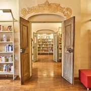 Stadtbibliothek Pirna Offene Holztüren führen in eine helle Bibliothek mit Bücherregalen und einem roten Sitzhocker auf einem Holzboden.