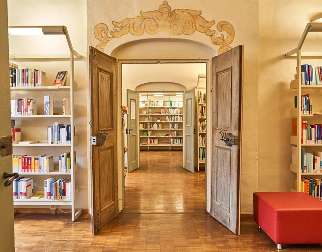 Stadtbibliothek Pirna Offene Holztüren führen in eine helle Bibliothek mit Bücherregalen und einem roten Sitzhocker auf einem Holzboden.