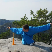 Taijiquan in der Sächsische-Schweiz Ein Mann in blauer Kleidung führt eine Kampfkunstpose mit einem Schwert auf einem Felsen aus, umgeben von grünen Bäumen und Bergen im Hintergrund.