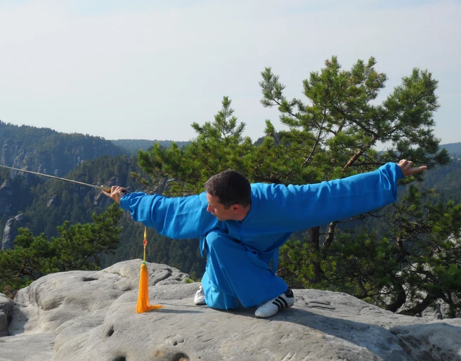 Taijiquan in der Sächsische-Schweiz Ein Mann in blauer Kleidung führt eine Kampfkunstpose mit einem Schwert auf einem Felsen aus, umgeben von grünen Bäumen und Bergen im Hintergrund.