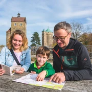 Familie beim Stadtspiel in Stolpen Eine Familie sitzt an einem Holztisch im Freien, betrachtet eine Karte und einen Reiseführer; im Hintergrund sind zwei historische Türme zu sehen.