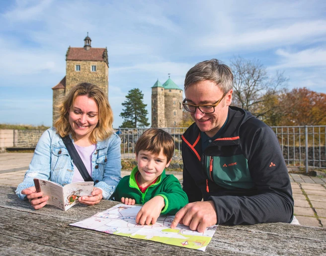 Familie beim Stadtspiel in Stolpen Eine Familie sitzt an einem Holztisch im Freien, betrachtet eine Karte und einen Reiseführer; im Hintergrund sind zwei historische Türme zu sehen.