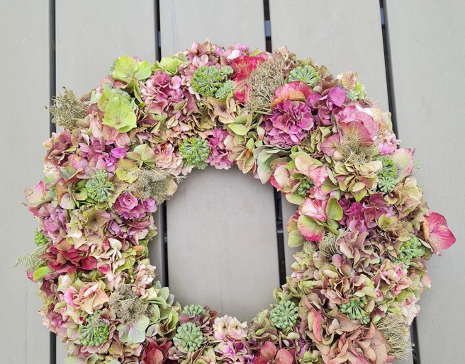 Hortensienkranz.jpg Ein Kranz aus getrockneten Hortensienblüten in Rosa- und Grüntönen liegt auf einem Holztisch.