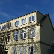Grundschule am Paulsberg  Ein dreistöckiges Schulgebäude mit beigen Wänden und weißen Fenstern, umgeben von Bäumen im Sonnenschein.