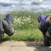 Zwei Personen in Wanderkleidung entspannen auf einem Weg, umgeben von grünem Gras und blühenden Blumen.