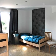 Helles Zimmer mit zwei Holzbetten, einem Stuhl und einem Fenster, das Licht auf den Holzboden wirft.