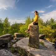 Lippischer Velmerstot-Kreis Lippe-Teutoburger-Wald-Tourismus-D-Ketz-001.jpg Frau sitzt auf historischem Grenzstein umgeben von Bäumen und bewaldeter Landschaft unter blauem Himmel.