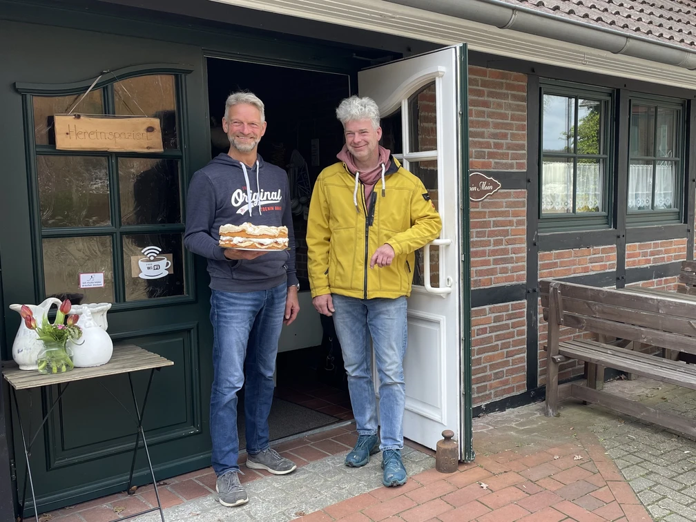 Eingangsbereich Hof Cafe Janßen Zwei freundliche Männer stehen vor einem Backsteinhaus; einer hält eine Torte, lächelnd in die Kamera.