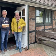 Eingangsbereich Hof Cafe Janßen Zwei freundliche Männer stehen vor einem Backsteinhaus; einer hält eine Torte, lächelnd in die Kamera.
