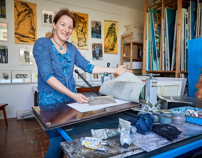 Künstlerin Anne Kern Eine Frau in einem blauen Kleid steht in einem Atelier vor einer Druckpresse, umgeben von Kunstwerken und Malutensilien.