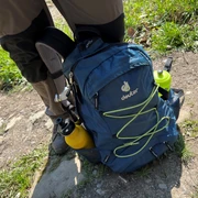Outdoor-Rucksack in Blau mit gelben Flaschen an den Seiten auf einem Wanderweg zwischen Gras und Steinen.