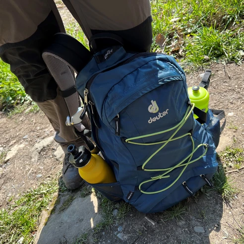 Rucksack Outdoor-Rucksack in Blau mit gelben Flaschen an den Seiten auf einem Wanderweg zwischen Gras und Steinen.