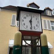 Spargeluhr Eine antike Standuhr mit einem großen Ziffernblatt steht vor einem pastellgelben Gebäude mit weißen Fensterläden.