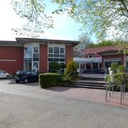 DRK_Silvana Dust_20230509_1980x1310.jpg Die Vorderseite des DRK-Krankenhauses zeigt den Haupteingang mit Türen. Vor dem Gebäude sind Gehwege, Parkplätze und einige Pflanzen