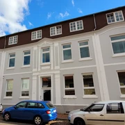 Stadtarchivar_Silvana Dust_20230510_1980x1310.jpg Ein Haus mit hellgrauem Anstrich und Seitenstreifen, davor parkende Autos