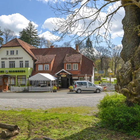 Forsthaus Blomberg mit rustikalem Fachwerk und grünem Schild, umgeben von Bäumen, ein weißes Auto davor.