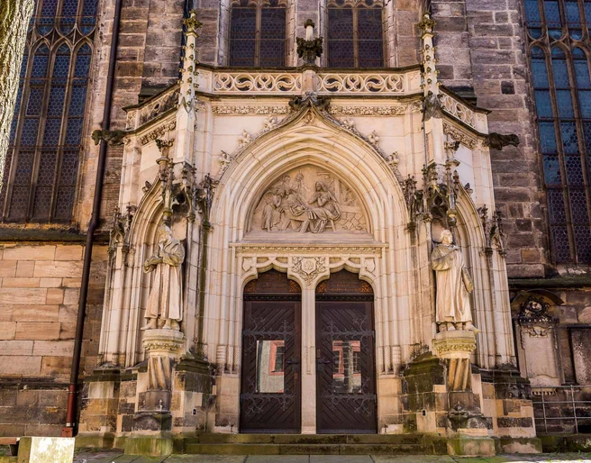 Tür der Stadtkirche St. Marien in Pirna Gotisches Kirchenportal mit zwei großen Holzflügeltüren, flankiert von steinernen Heiligenstatuen und einem Relief über dem Eingang.
