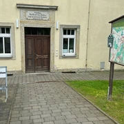 Eingang zur Touristinformation Rosenthal-Bielatal in der Gemeindeverwaltung  Eingang eines historischen Gebäudes mit der Aufschrift "Die Kraft des Volkes liegt in unseren Kindern", daneben eine Infotafel und ein Schild "Touristinformation".