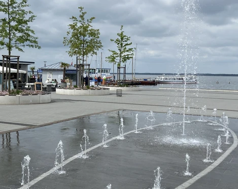 Wasserspiele mit Fontänen auf einer Terrasse am Wasser, umgeben von Bäumen und grauem Himmel.