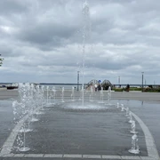 Springbrunnen und Strandterrassen am Seeufer, im Hintergrund bewölkter Himmel.