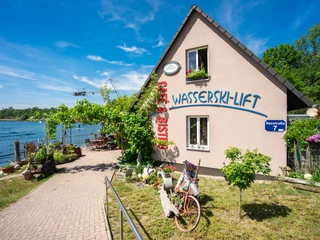 Wasserskilift, Gastonomie und Café am Kulkwitzer See Ein idyllisches Haus mit großer Terrasse am Kulkwitzer See, umgeben von blühenden Pflanzen. Der Wasserskilift im Hintergrund bietet sportliche Möglichkeiten in malerischer Kulisse. Ein Fahrrad mit Blumenschmuck verleiht dem Ambiente eine gemütliche Note.