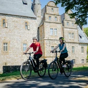 Die Schelenburg bei Bissendorf Zwei Radfahrer vor einem historischen steinernen Herrenhaus bei sonnigem Wetter.