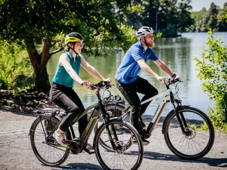 Rubbenbruchsee Zwei Radfahrer mit Schutzhelmen fahren fröhlich auf einem sonnigen Weg entlang eines Sees.