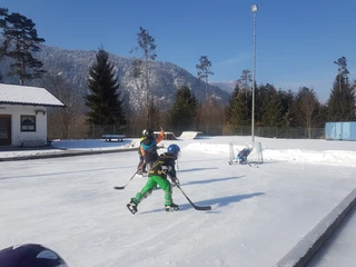 Eisplatz Farchant  2 Eishockeyspielende Kinder mit Tor, im Hintergrund Bäume und der Umkleideraum