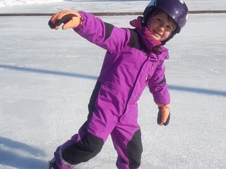 Eisplatz Farchant  kleines Mädchen im lila Schneeanzug und lila Helm auf Schlittschuhen auf dem Eisplatz, im Hintergrund Zaun und Bäume