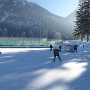 Eisplatz Farchant  5 Personen auf dem im Schatten liegendem Eisplatz. Im Hintergrund das Tor, Zaun, Bäume, Flutlichtlampen sowie der Hohe Fricken