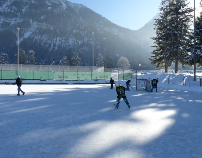 Eisplatz Farchant  5 Personen auf dem im Schatten liegendem Eisplatz. Im Hintergrund das Tor, Zaun, Bäume, Flutlichtlampen sowie der Hohe Fricken
