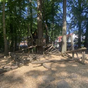 Spielplatz an der Grundschule Spielstruktur aus Holz in einem schattigen Waldabschnitt, umgeben von Bäumen und weichem Bodenbelag.
