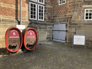Zwei rote Eingangstüren in großen Holzfässern, nebeneinander vor einem historischen Backsteingebäude.