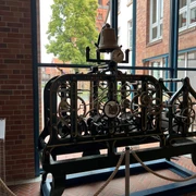 Historisches Uhrwerk mit Zahnrädern, umgeben von zwei Ziegelwänden und großen Fenstern.