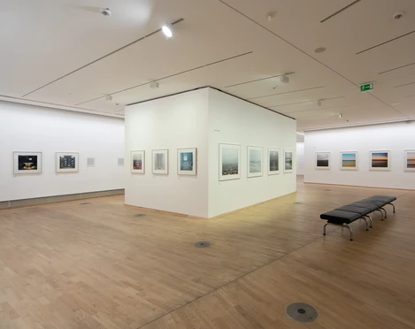 The Photographic Collection/SK Stiftung Kultur Ein lichtdurchfluteter Ausstellungsraum in einer Galerie mit Holzfußboden, an dessen Wänden gerahmte Fotografien hängen.A light-flooded exhibition space in a gallery with a wooden floor and framed photographs hanging on the walls.