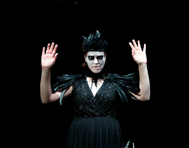 Theater im Bauturm Eine Frau in schwarzem Kleid mit Federdetails trägt dramatisches Make-up und hebt die Hände.A woman in a black dress with feather details wears dramatic make-up and raises her hands.