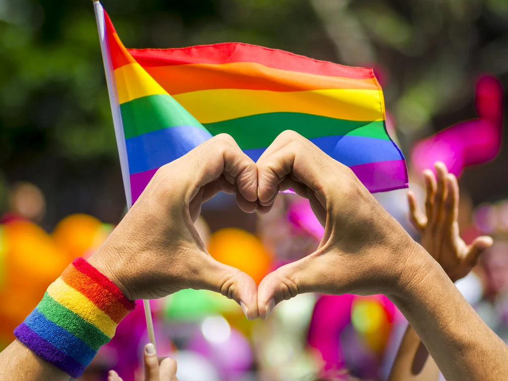 LGBTQ+ heart Hände formen ein Herz vor einer RegenbogenflaggeHands form a heart in front of a rainbow flag